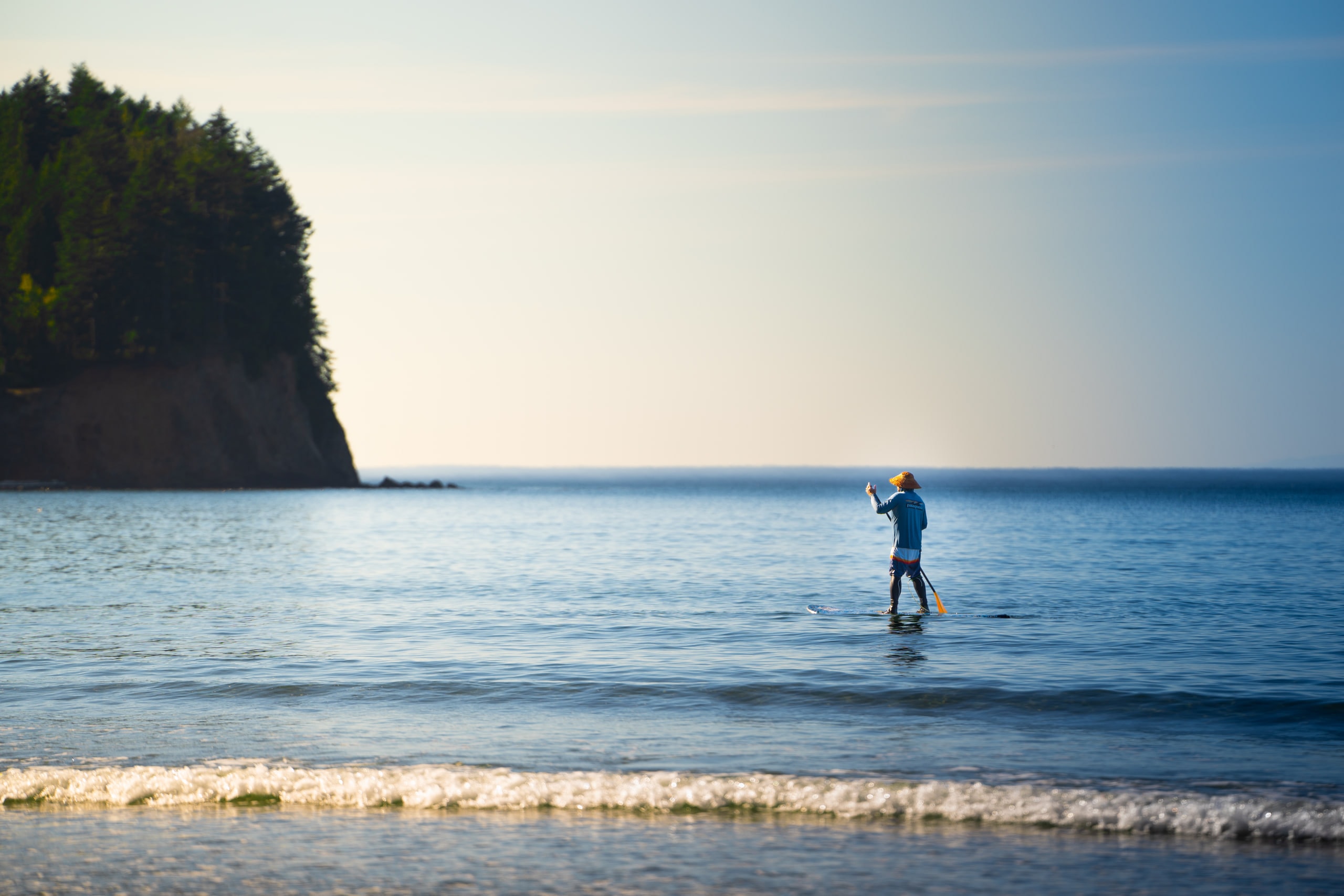 A paddleboarder crossing flat water beneath a headland at dusk