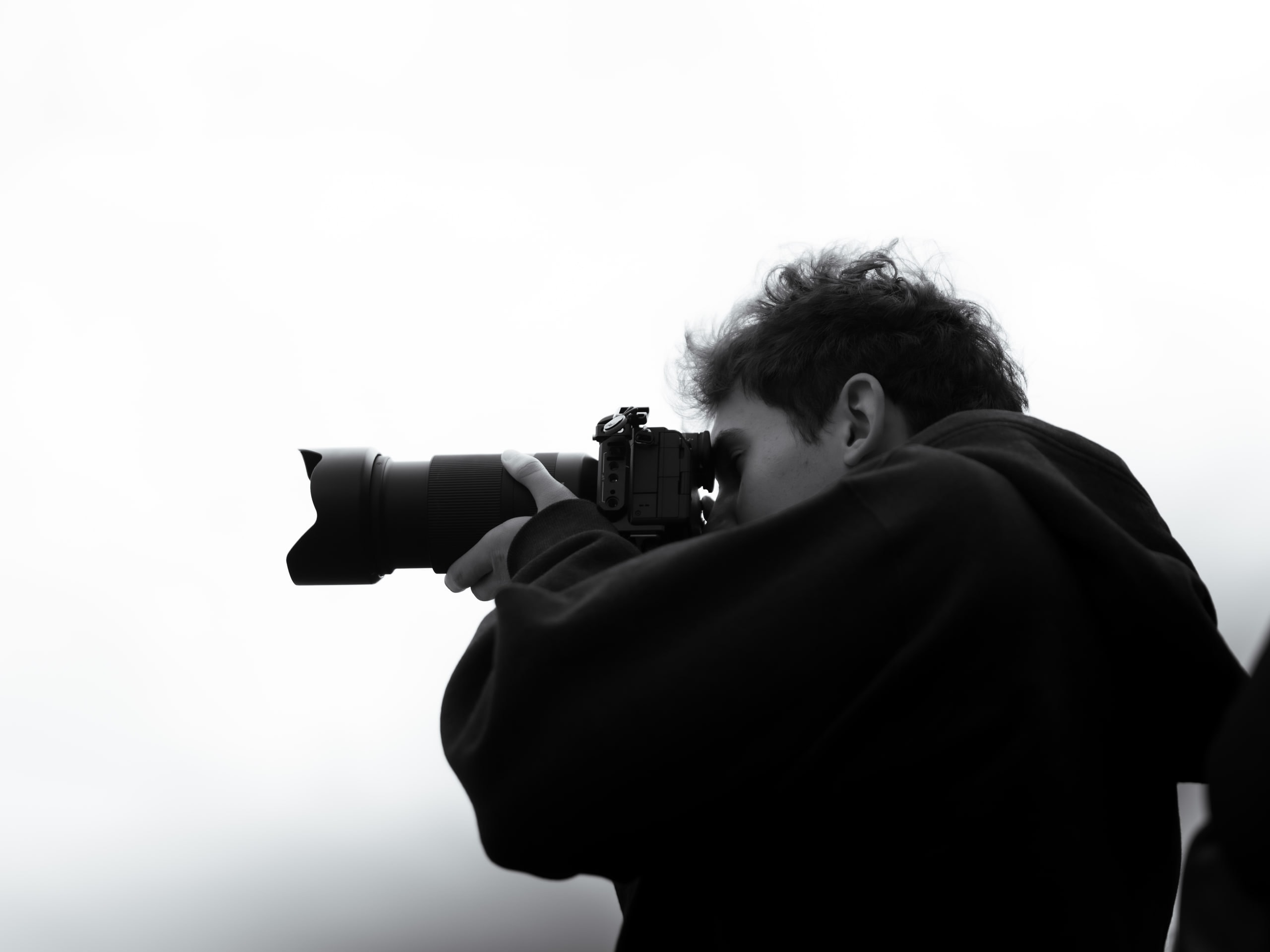 The photographer in profile, hooded, shooting with a long telephoto lens, in black and white